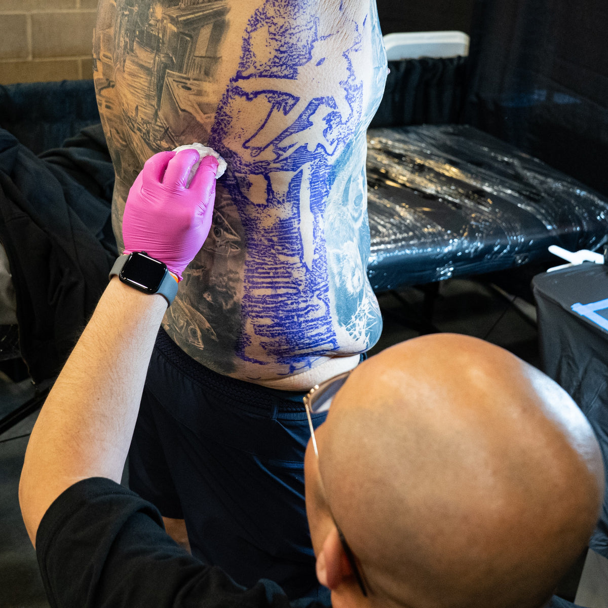 A tattoo artist wearing a black shirt and pink gloves applies a blue-purple tattoo design onto a client's upper back in a tattoo studio.