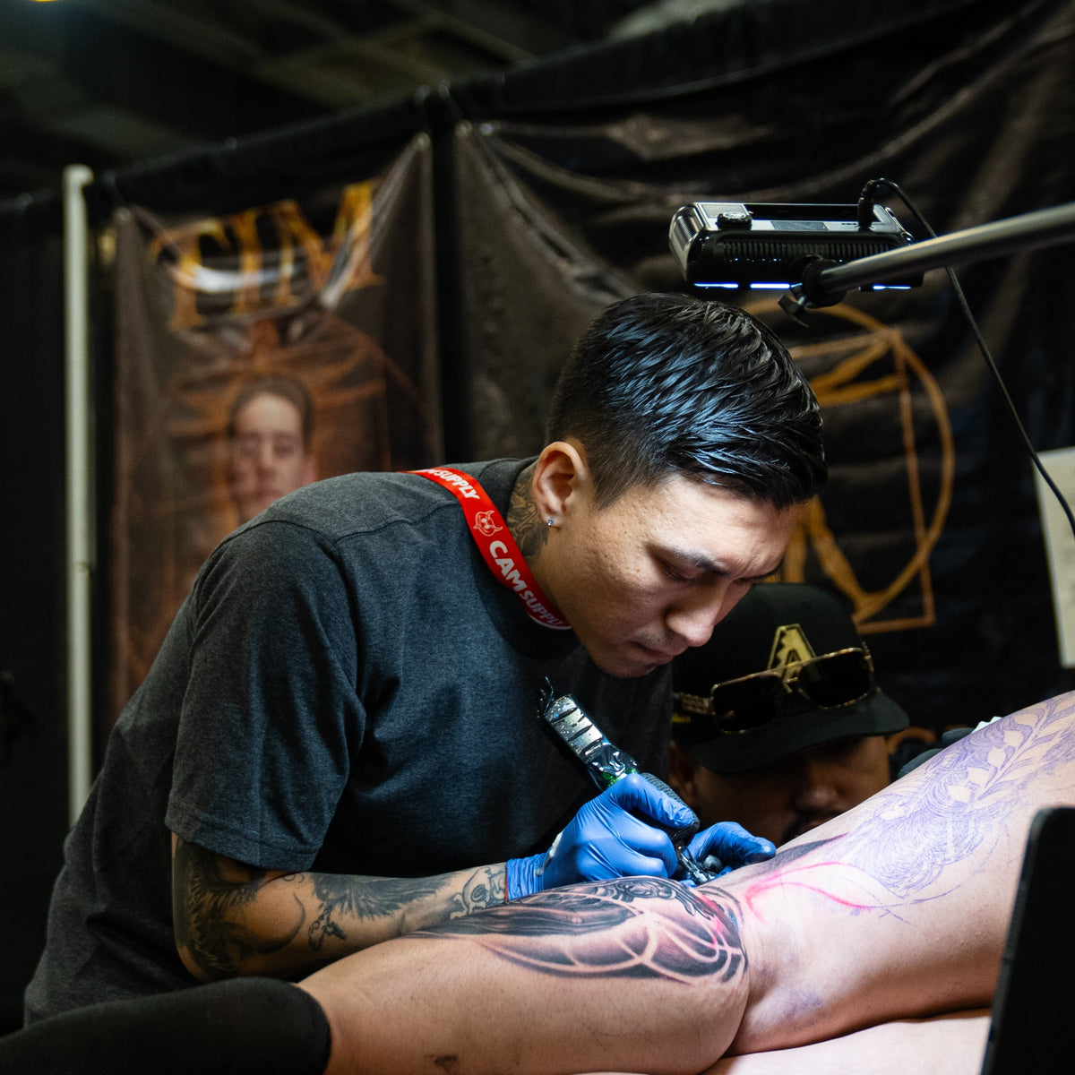 A tattoo artist wearing blue gloves and a black shirt applies a tattoo to a person's upper leg at a tattoo studio. 