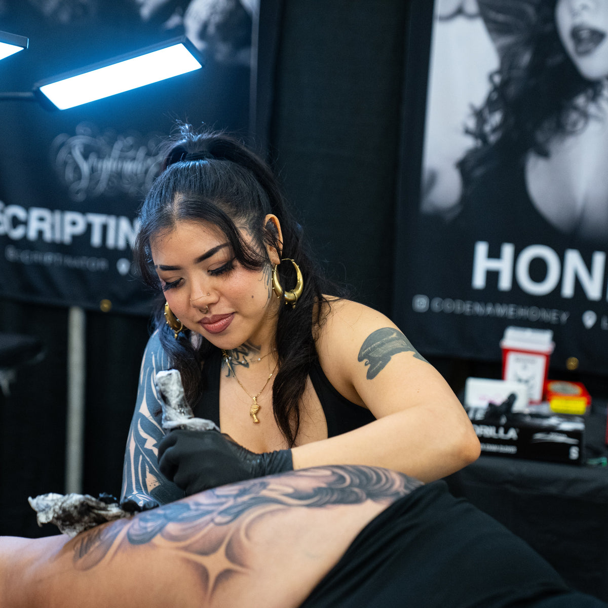 A tattoo artist wearing black gloves and gold hoop earrings applies a tattoo to a client's upper back. The artist has dark hair in a ponytail, a nose ring, and visible arm tattoos.