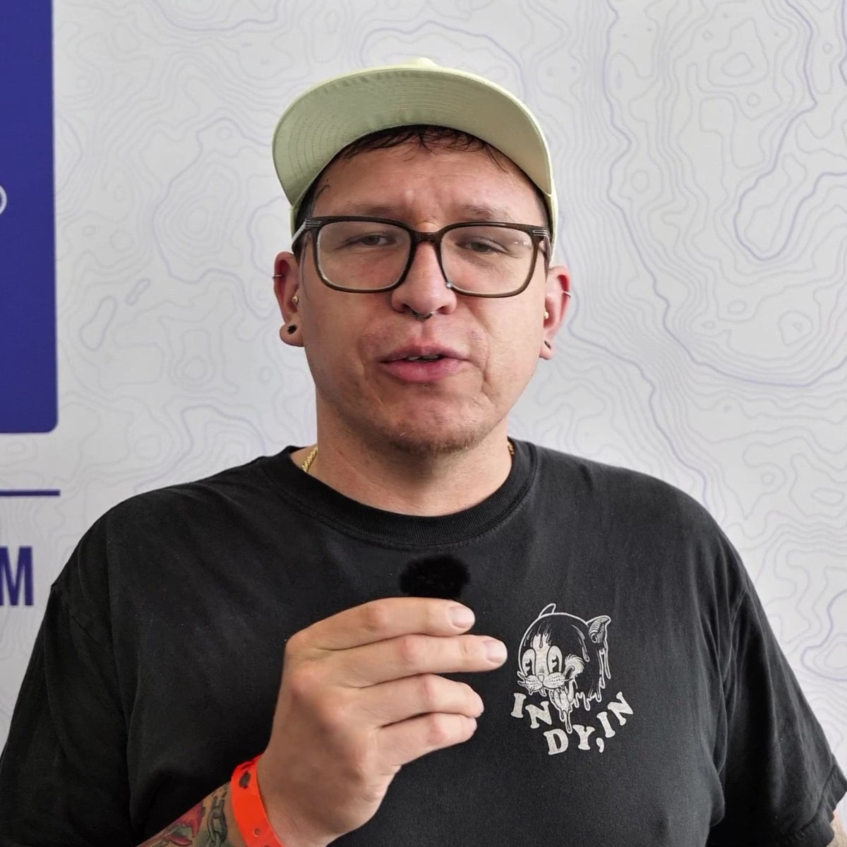 A man wearing a light green cap, black glasses, and a black t-shirt with a white graphic print that reads 'IN DYIN', holding a small black microphone in front of his chest. The background is a white wall with a faint topographic line pattern.