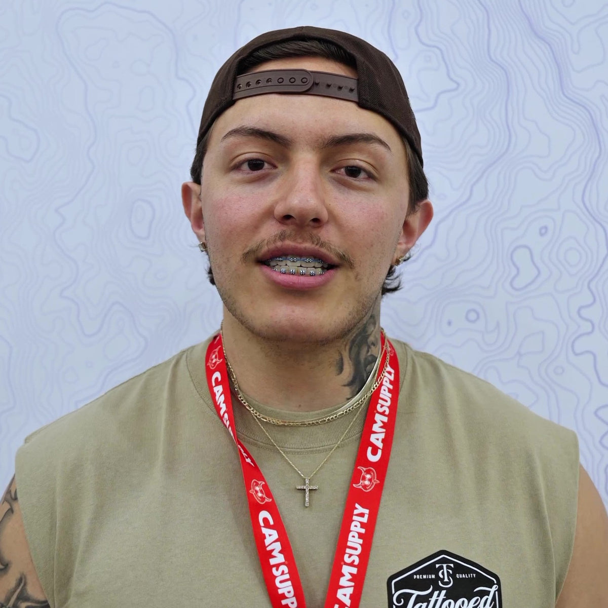A man wearing a brown cap backwards, a beige sleeveless shirt, and a red 'Cam Supply' lanyard, with a black 'Cam Tattooed' patch on his chest, set against a light blue patterned background.