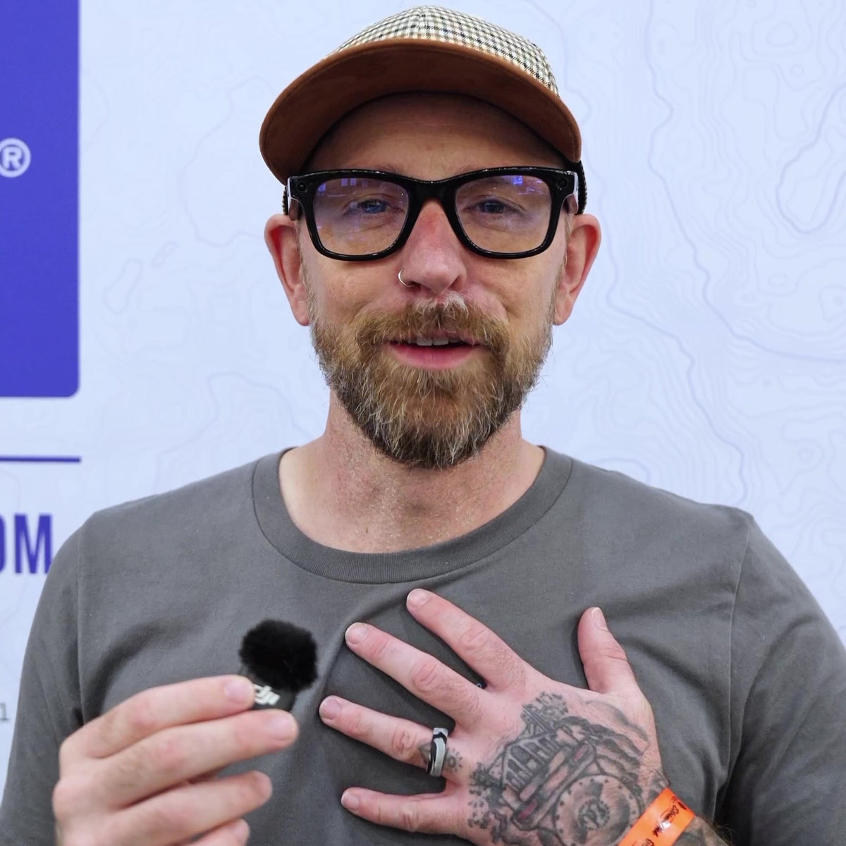 A man with a beard, black-rimmed glasses, and a brown cap holds a small silver ring with a black pom-pom clip in front of his chest, wearing a gray t-shirt and a tattooed hand.